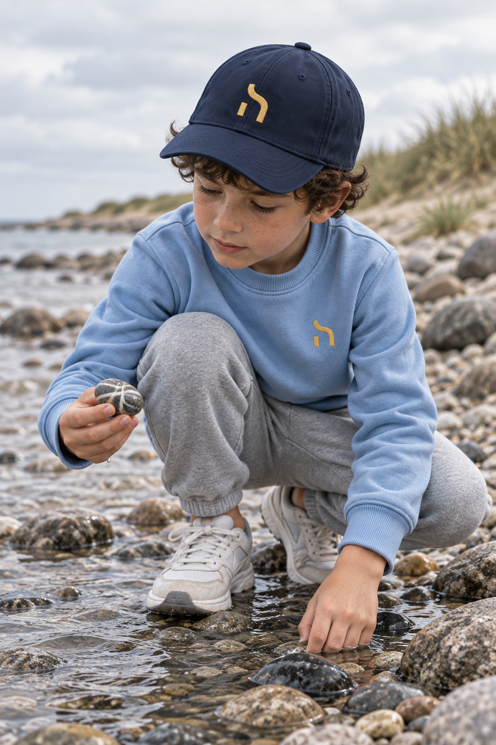 Junge trägt eine dunkelblaue Haensch Coast Kids Cap und ein hellblaues Haensch-Sweatshirt an einem steinigen Ostseestrand.