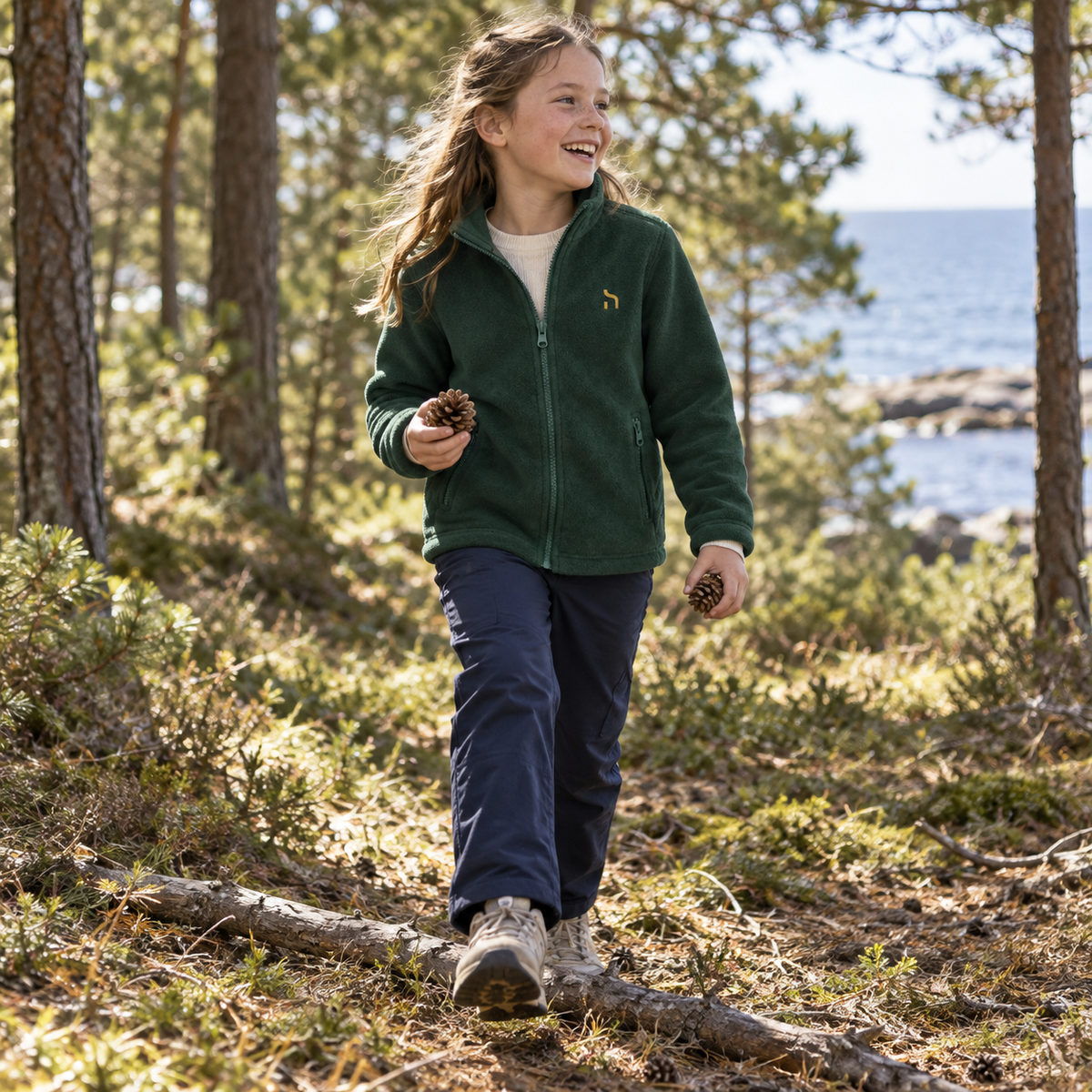 Mädchen trägt die flaschengrüne Haensch Coast Kids Fleecejacke mit goldenem Brustzeichen und sammelt Zapfen in einem Küstenwald an der Ostsee.
