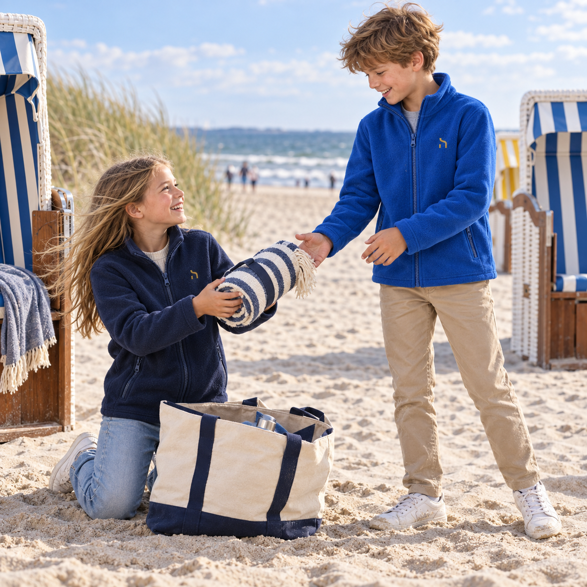 Zwei Kinder tragen Haensch Coast Kids Fleecejacken in Deep Navy und Royal Blue und packen gemeinsam eine Decke an einem Ostseestrand aus.