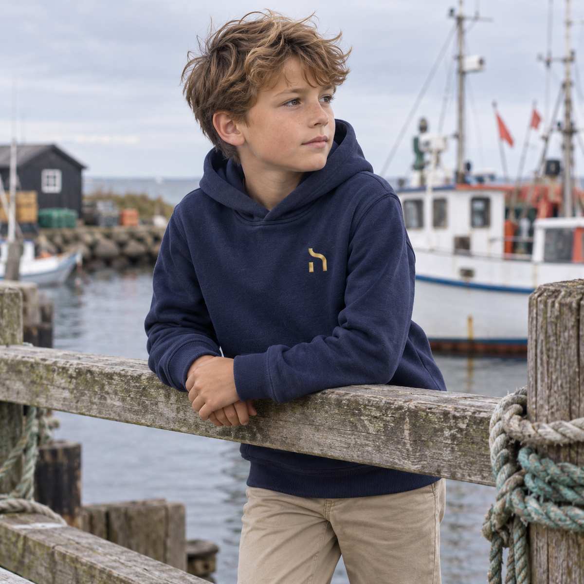 Junge trägt den marineblauen Haensch Coast Kids Hoodie mit goldenem Haensch-Zeichen an einem kleinen Ostseehafen.