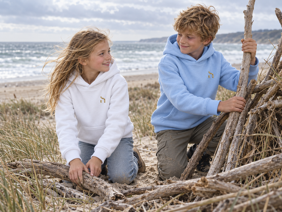 Mädchen und Junge tragen Haensch Coast Kids Hoodies in Weiß und Himmelblau beim Bauen mit Treibholz am Ostseestrand.
