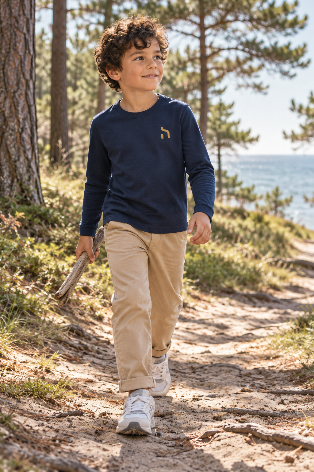 Junge trägt das marineblaue Haensch Coast Kids Longsleeve mit goldenem Brustzeichen auf einem sandigen Weg im Küstenwald.