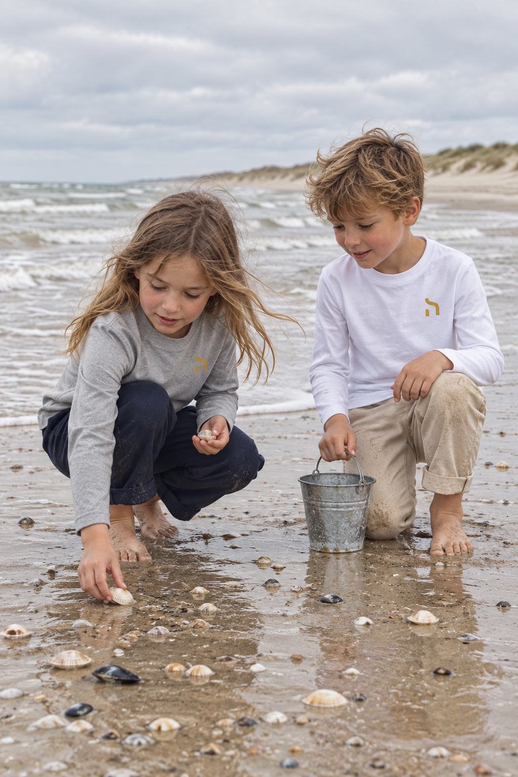Zwei Kinder tragen Haensch Coast Kids Longsleeves in Heather Grey und Weiß und suchen gemeinsam Muscheln am Ostseestrand.