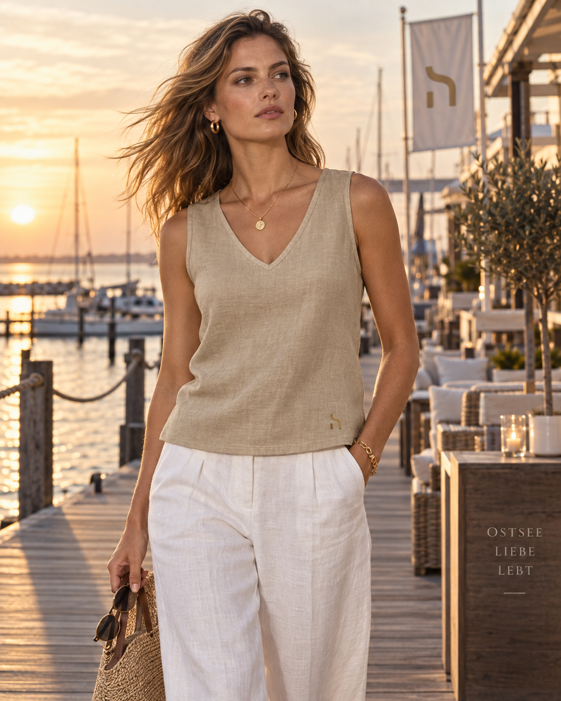 Frau im Haensch Coast Linen V-Top Damen in Coast Sand Khaki mit goldenem H-Logo bei einem hochwertigen sommerlichen Küsten-Lifestyle-Look