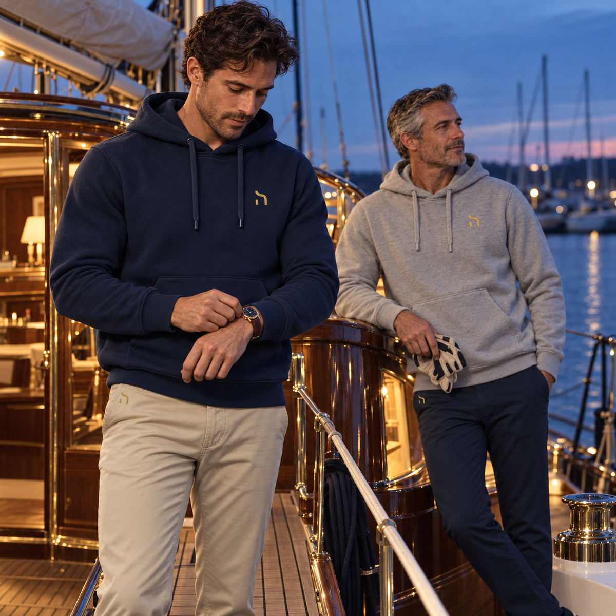 Zwei Männer tragen Haensch Coast Premium Hoodies in Deep Navy und Heather Grey auf einer klassischen Segelyacht im abendlichen Yachthafen.
