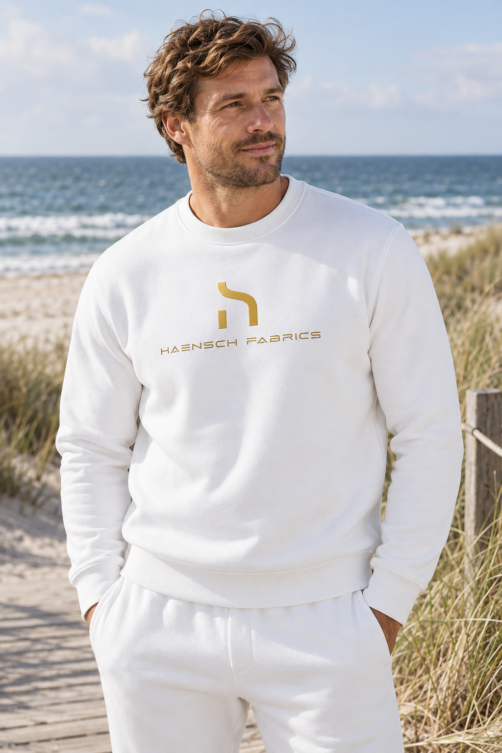 Männliches Model trägt den weißen Haensch Coast Signature Crewneck mit goldenem Haensch Fabrics Logo vor Dünen und Ostsee