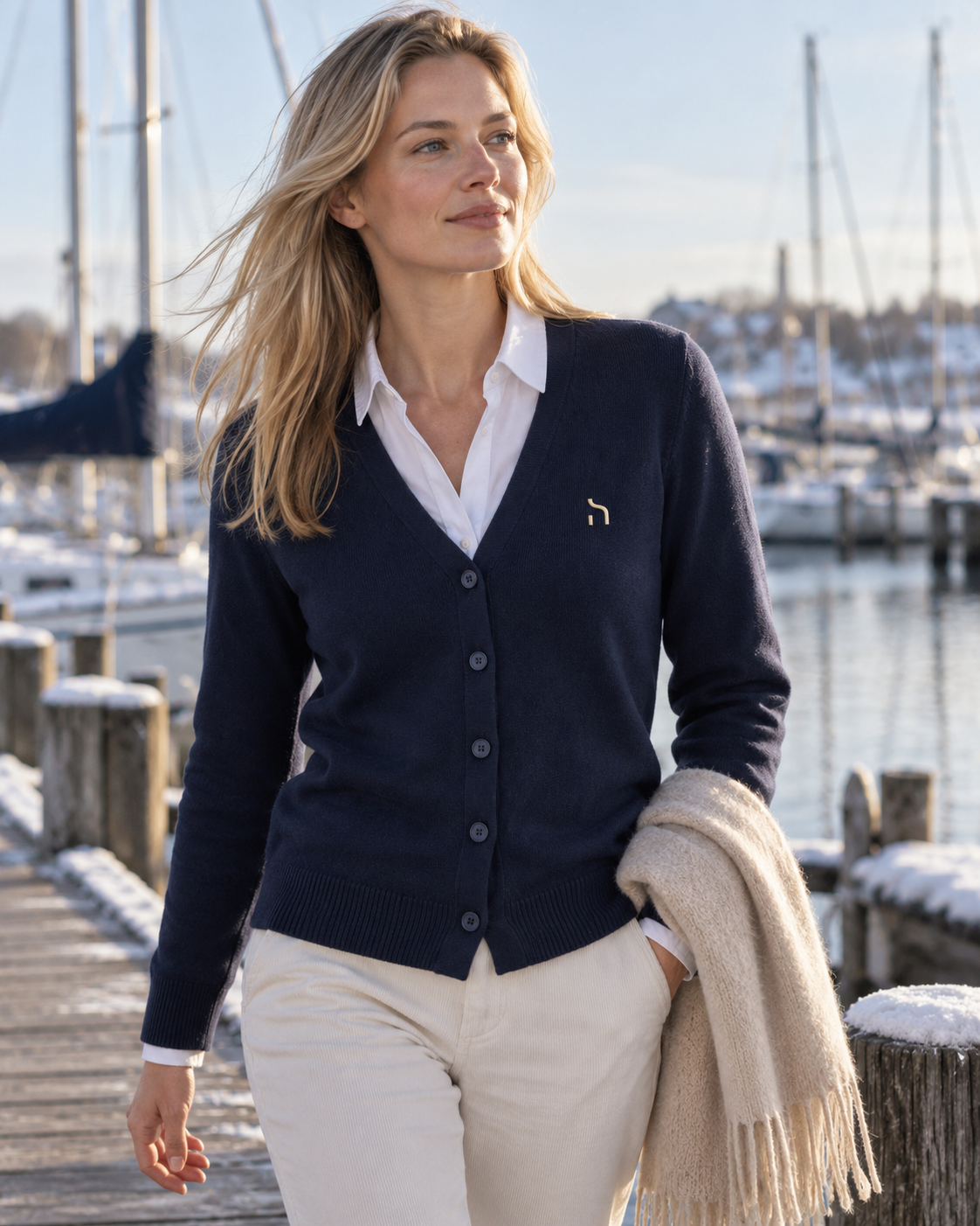 Weibliches Model trägt den Haensch Heritage Supima Cardigan Damen in Deep Navy über weißer Bluse mit goldenem Hänsch-H-Signet in einer winterlichen Hafen-Kulisse