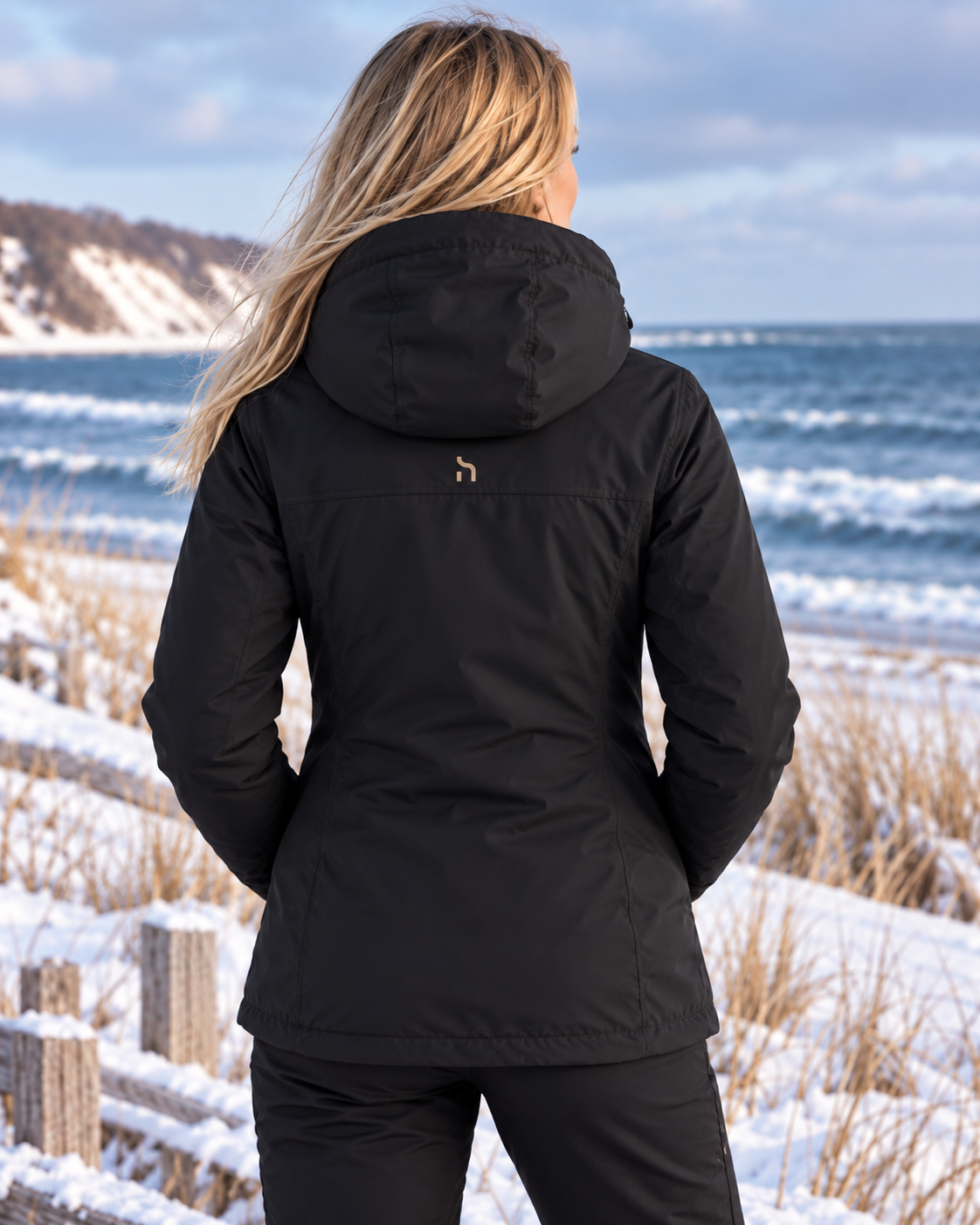 Rückansicht der Haensch Northern All Weather Winter Jacket Damen in Winter Black mit kleinem goldfarbenem Haensch-h mittig unter der Kapuze vor winterlicher Küstenkulisse.