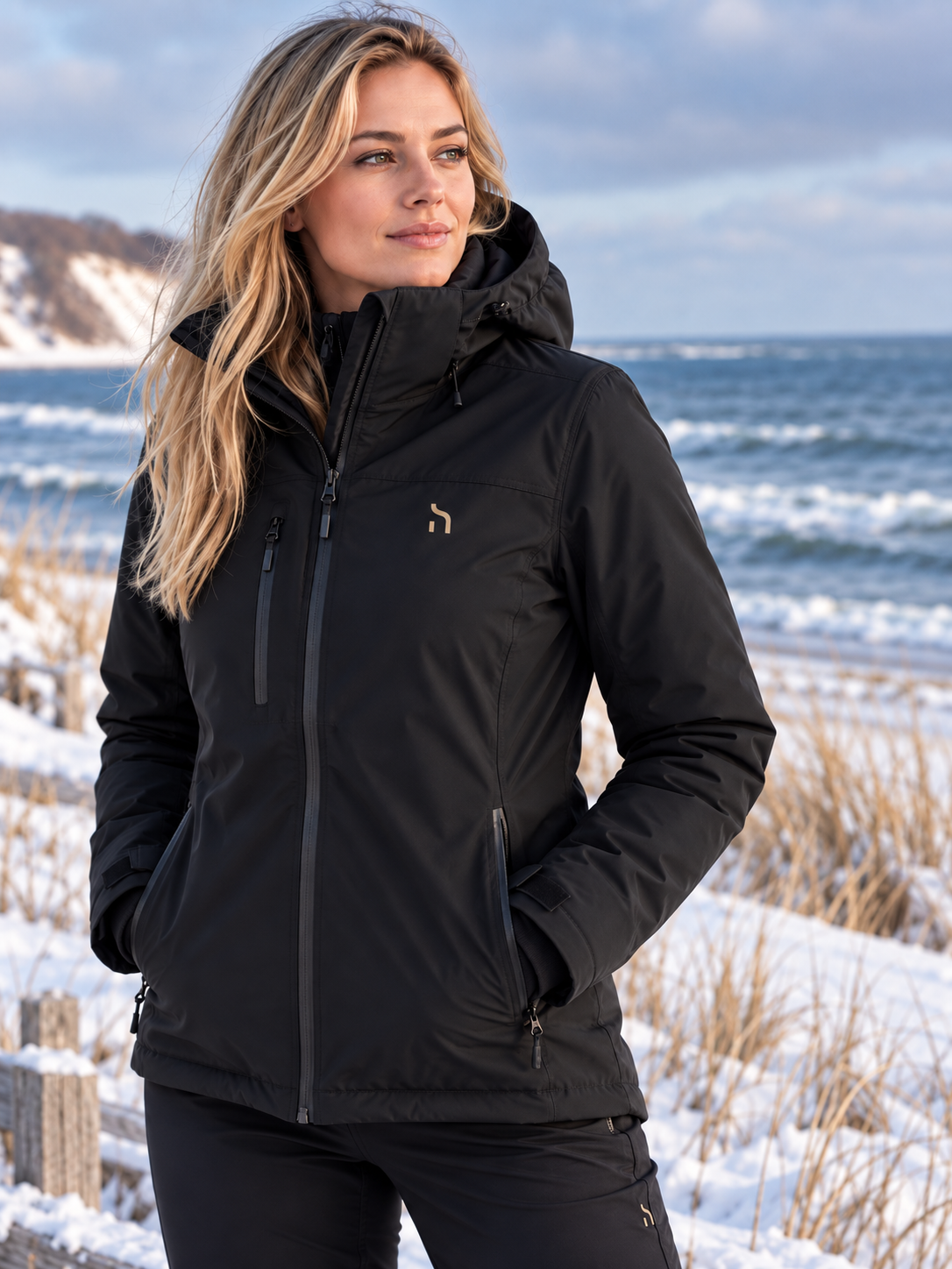 Vorderansicht der Haensch Northern All Weather Winter Jacket Damen in Winter Black mit kleinem goldfarbenem Haensch-h auf der Brust vor winterlicher Küstenkulisse.