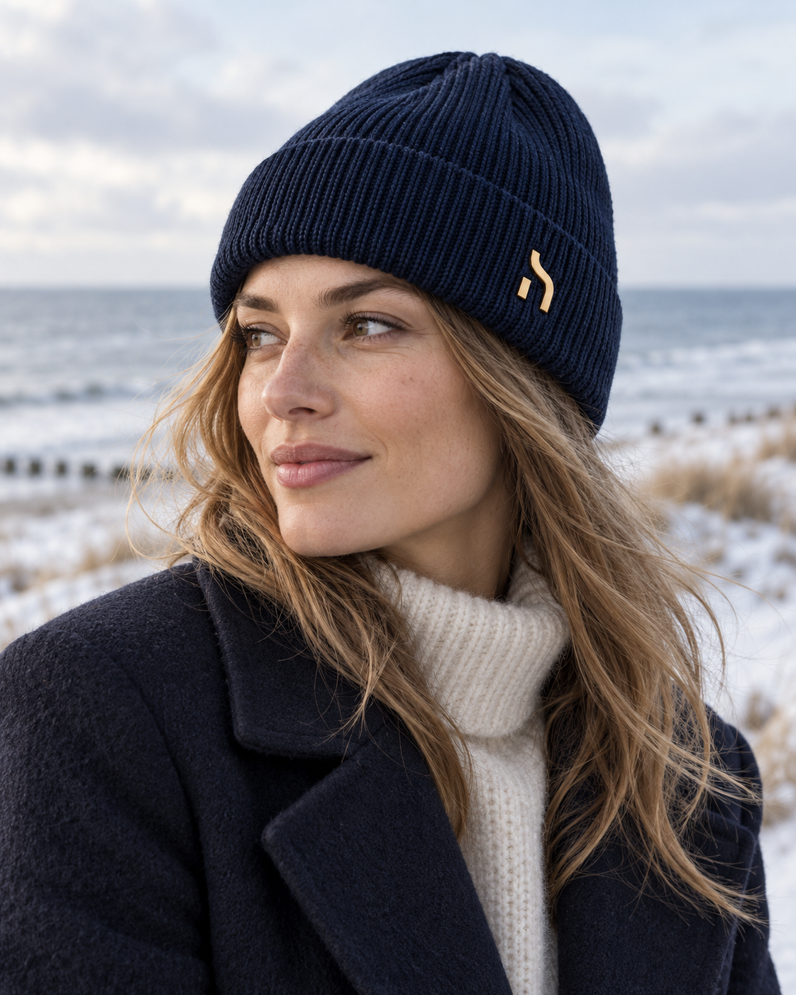 Weibliches Model trägt die Haensch Northern Cuffed Beanie in Deep Navy mit goldenem Hänsch-H-Signet vor winterlicher Ostsee-Kulisse