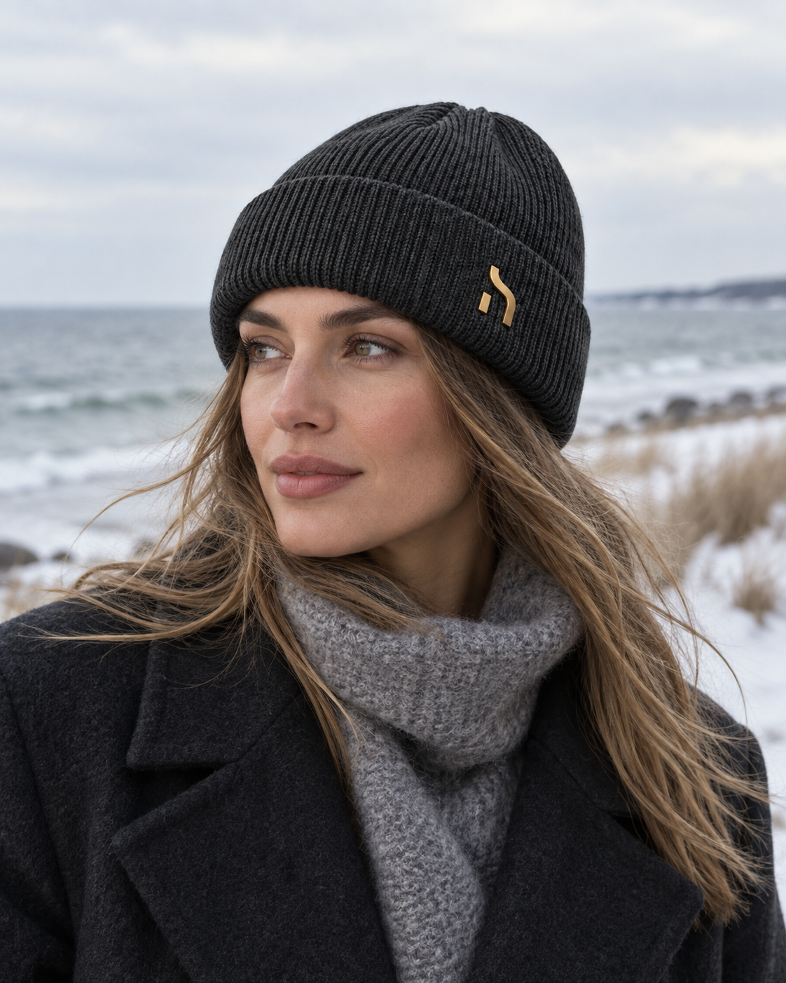 Weibliches Model trägt eine graue Haensch Northern Cuffed Beanie mit goldenem Hänsch-H-Signet zu elegantem Wintermantel an der Ostsee