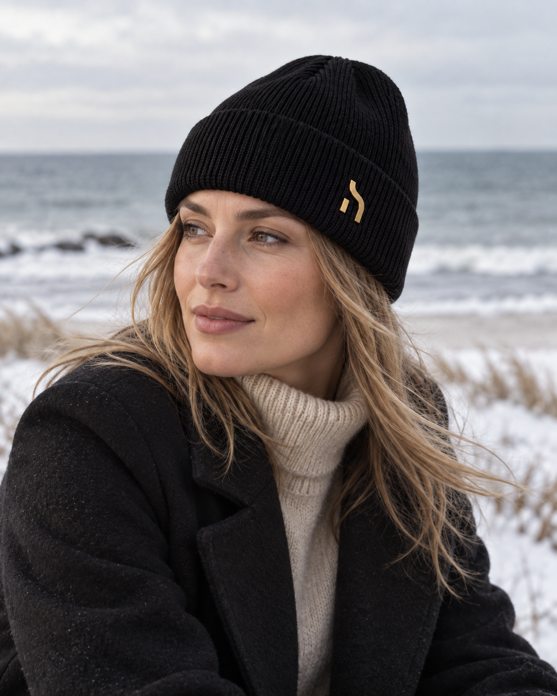 Weibliches Model trägt die Haensch Northern Cuffed Beanie in Winter Black mit goldenem Hänsch-H-Signet an der winterlichen Ostseeküste
