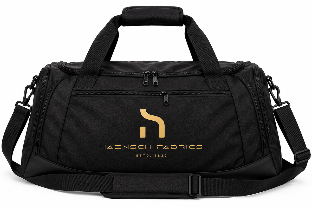 Black Haensch Signature Duffle Bag with gold Haensch Fabrics logo, dual carry handles, front zip pocket and adjustable shoulder strap on a white background.