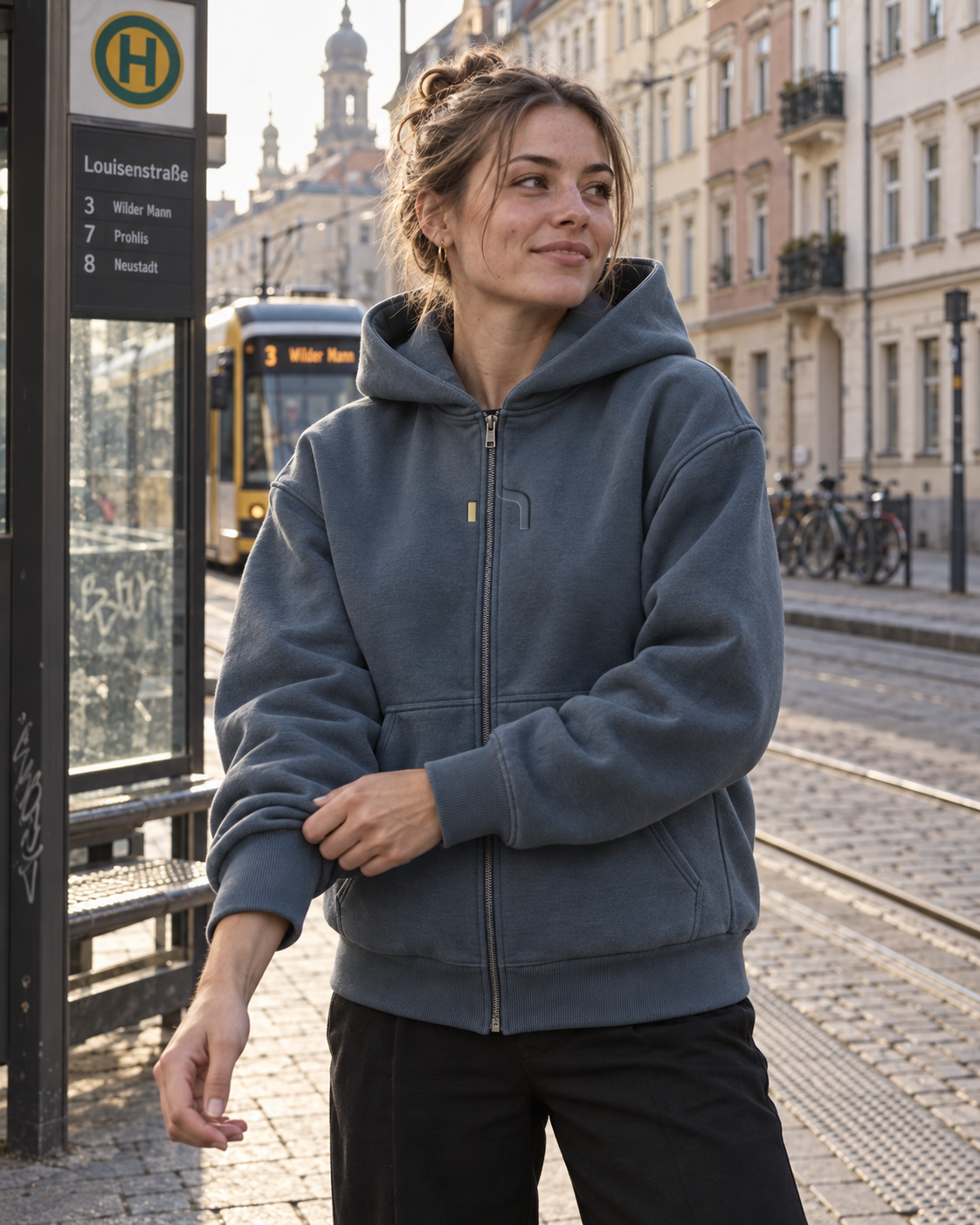 Junge Frau trägt den Haensch Studio Ultra Heavy Zip Hoodie in Vintage Blue an einer Straßenbahnhaltestelle in Dresden.