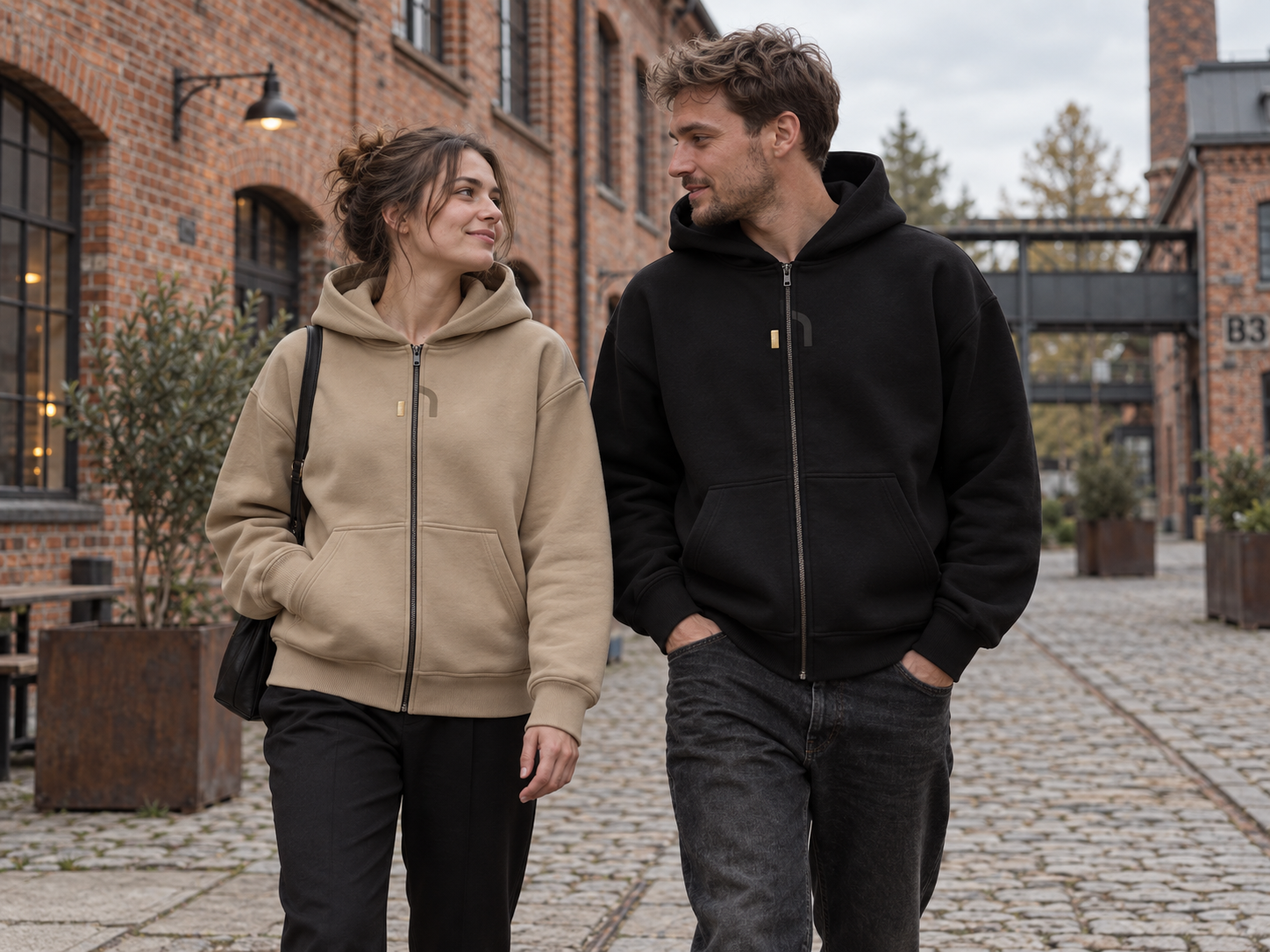 Junges Paar trägt Haensch Studio Ultra Heavy Zip Hoodies in Union Beige und Schwarz in einem sächsischen Industriehof.