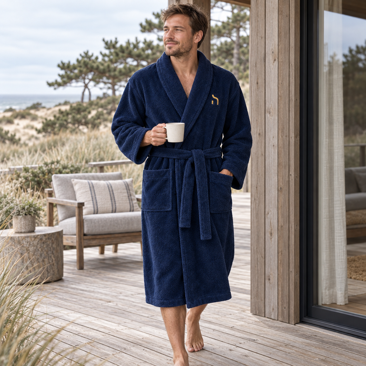 Junger Mann traegt den dunkelblauen Haensch Coast Classic Bathrobe mit goldenem Haensch-Zeichen auf der Terrasse eines Hauses in den Ostseeduenen.