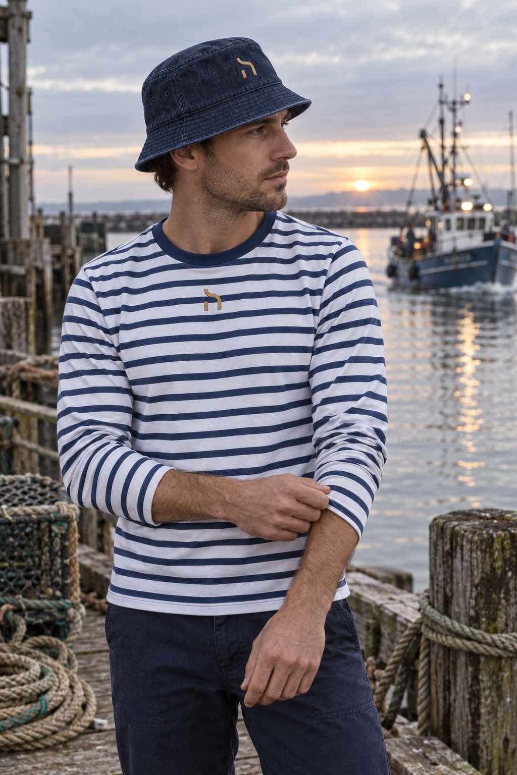 Junger Mann traegt den marineblauen Haensch Coast Denim Bucket Hat und ein gestreiftes Haensch Coast Longsleeve in einem kleinen Fischereihafen.