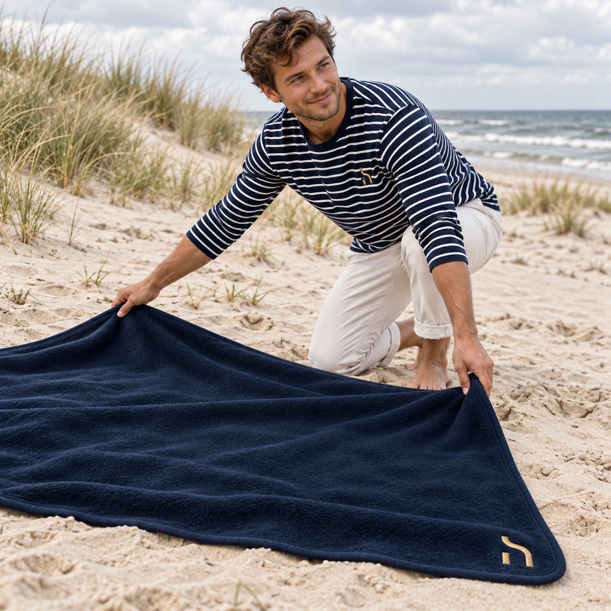 Mann breitet die navyfarbene Haensch Coast Fleece Picknickdecke mit goldenem Haensch-Zeichen am Ostseestrand aus.