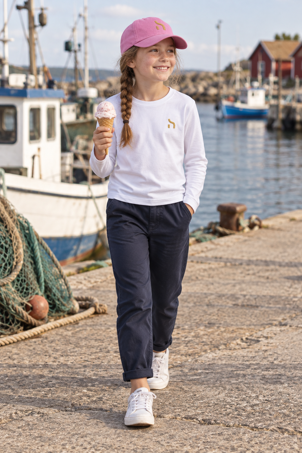 Mädchen trägt eine rosafarbene Haensch Coast Kids Cap und ein weißes Haensch-Longsleeve an einem kleinen Ostseehafen.