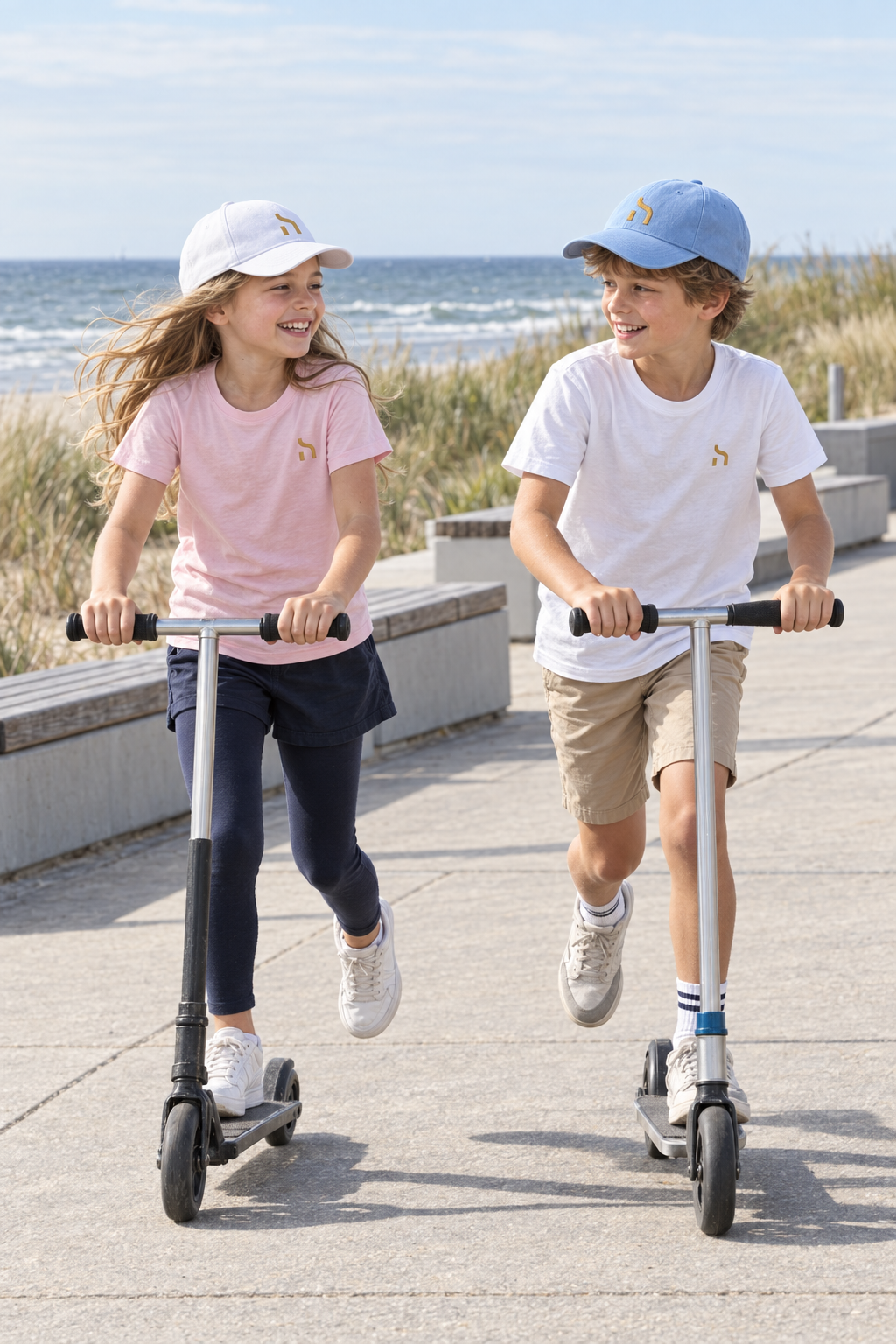 Zwei Kinder fahren mit Tretrollern über eine Ostsee-Promenade und tragen Haensch Coast Kids Caps in Weiß und Sky Blue sowie passende Haensch-Shirts.