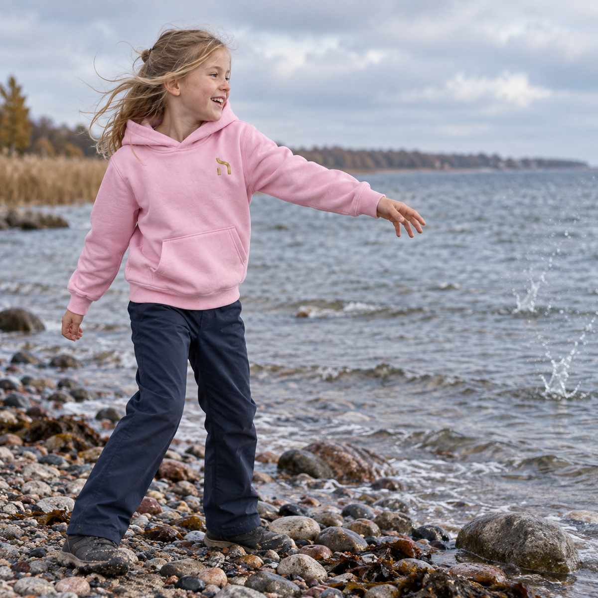 Mädchen trägt den hellrosa Haensch Coast Kids Hoodie mit goldenem Haensch-Zeichen und wirft einen Stein ins Wasser.