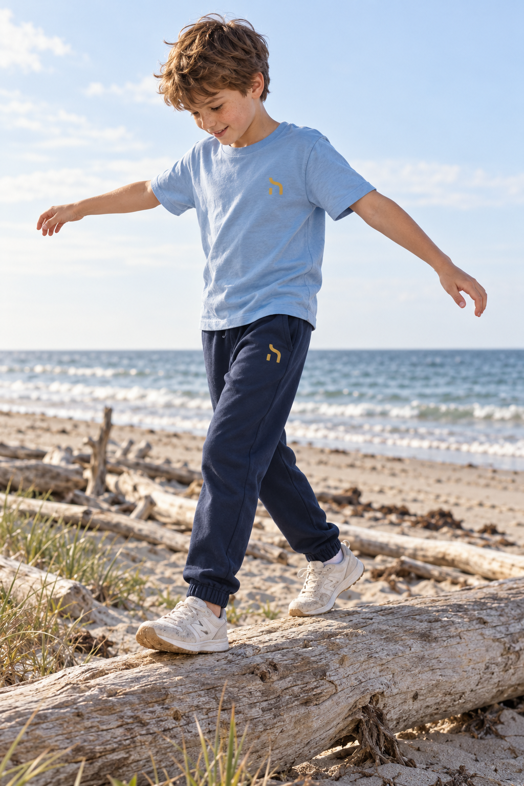 Junge trägt die marineblaue Haensch Coast Kids Jogginghose und ein hellblaues Haensch T-Shirt beim Balancieren auf Treibholz.