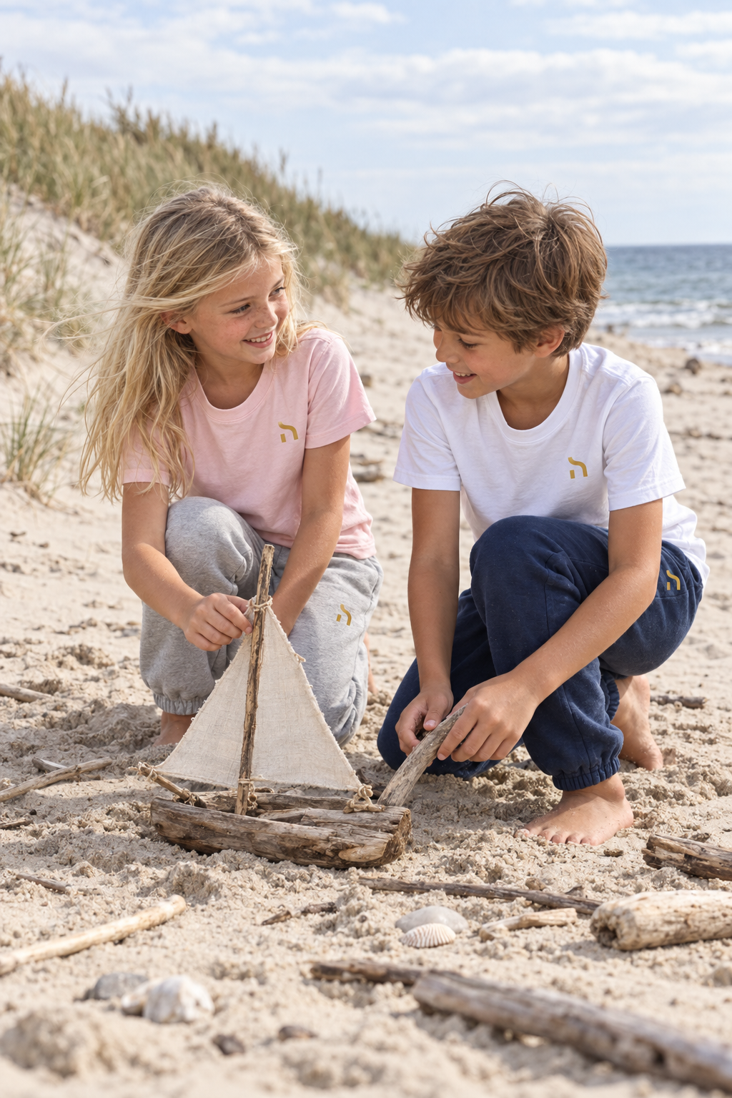 Zwei Kinder tragen Haensch Coast Kids Jogginghosen in Heather Grey und Deep Navy und bauen am Ostseestrand ein kleines Segelboot.