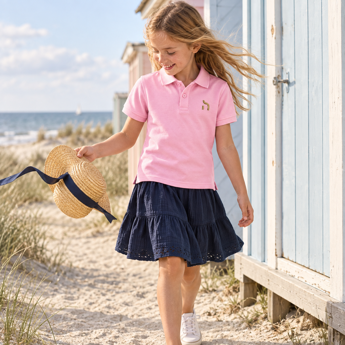 Mädchen trägt das hellrosa Haensch Coast Kids Poloshirt mit goldenem Haensch-Zeichen und hält einen Sonnenhut am Ostseestrand.