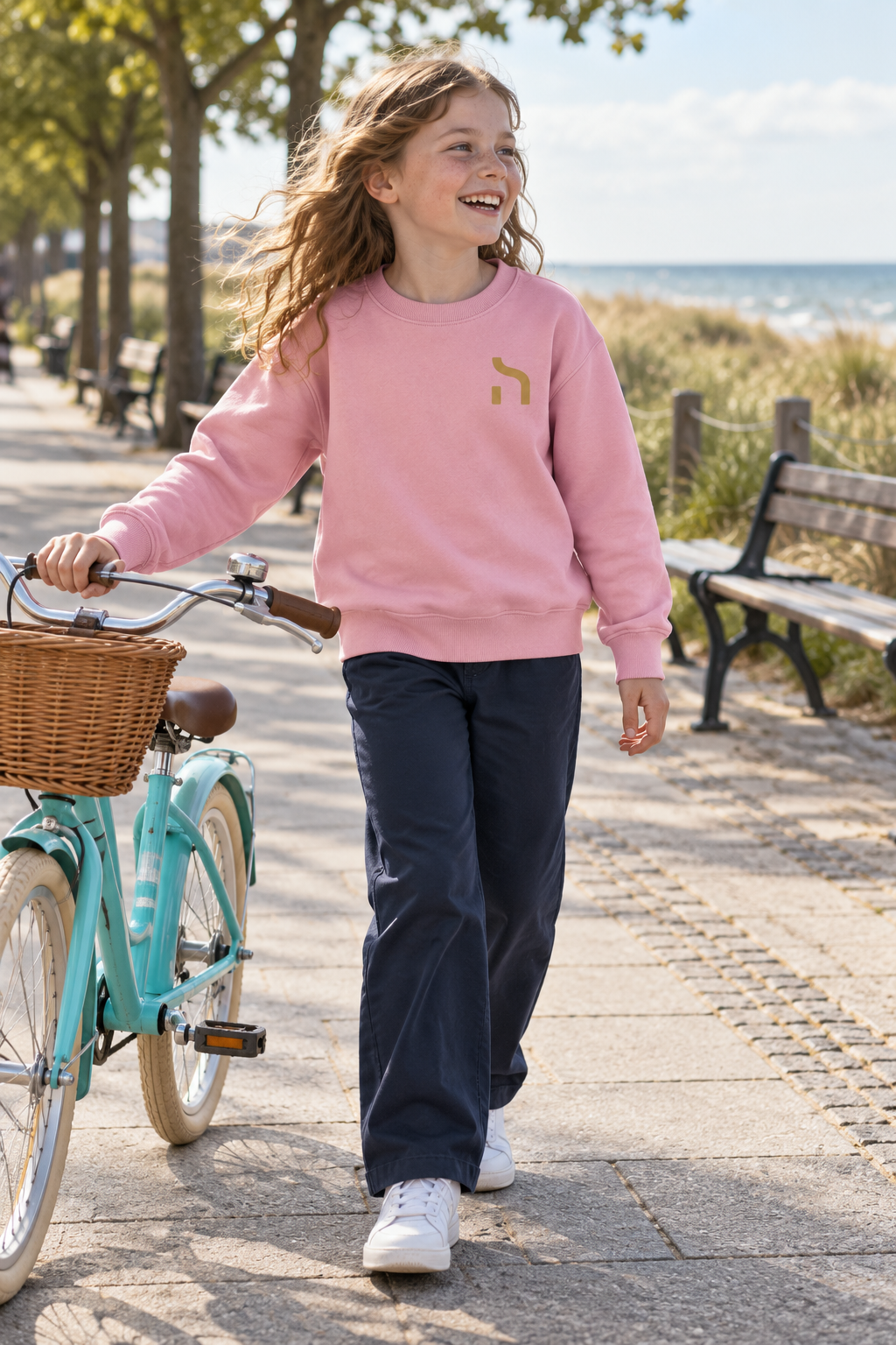 Mädchen trägt das rosafarbene Haensch Coast Kids Sweatshirt mit goldenem Brustzeichen und schiebt ein Fahrrad an der Strandpromenade.
