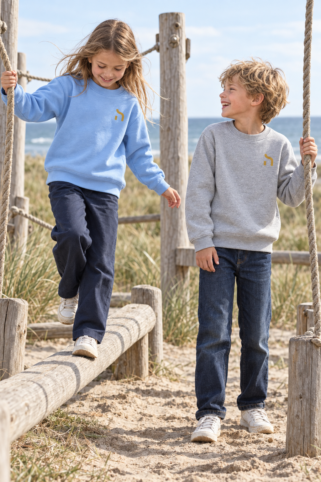 Zwei Kinder tragen Haensch Coast Kids Sweatshirts in Sky Blue und Heather Grey auf einem Spielplatz an der Ostseeküste.