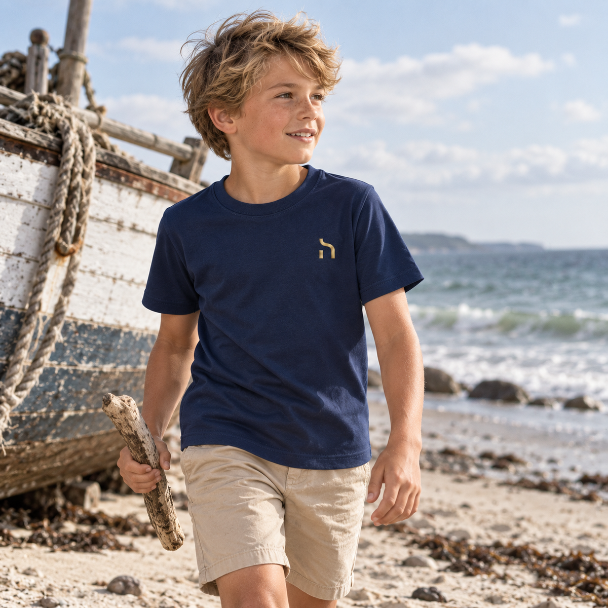 Junge trägt das marineblaue Haensch Coast Kids T-Shirt mit goldenem Haensch-Zeichen an einem Fischerboot am Ostseestrand.