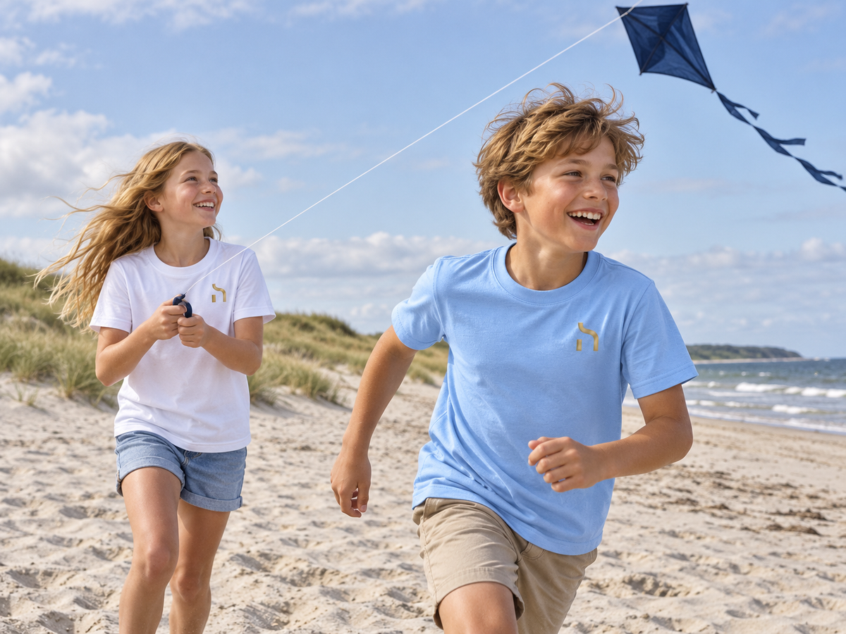 Mädchen und Junge tragen Haensch Coast Kids T-Shirts in Weiß und Himmelblau beim Drachensteigen am Ostseestrand.