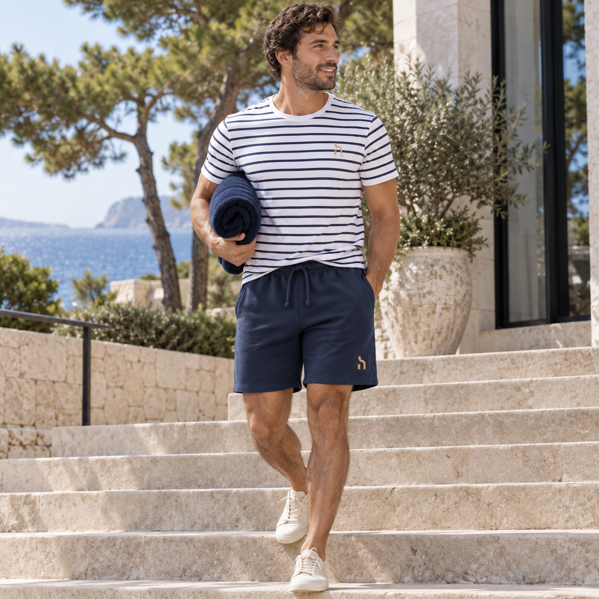 Mann traegt die Deep-Navy-farbenen Haensch Coast Lightweight Shorts mit gestreiftem Haensch Coast T-Shirt an einem modernen Strandhotel.