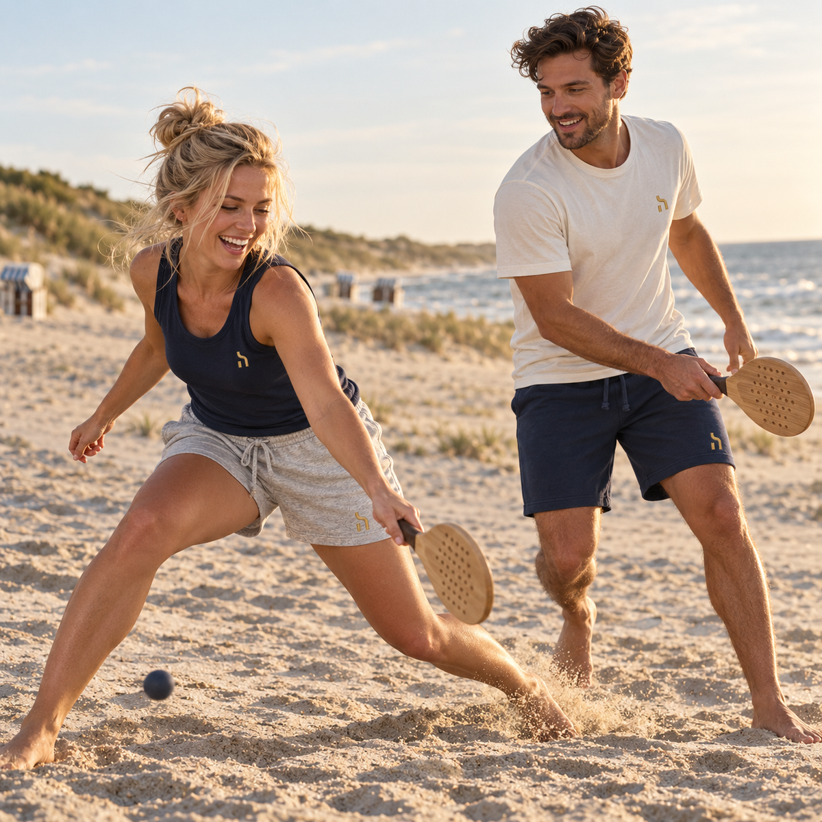 Junges Paar spielt am Ostseestrand in Haensch Coast Lightweight Shorts in Heather Grey und Deep Navy sowie passenden Haensch Oberteilen.