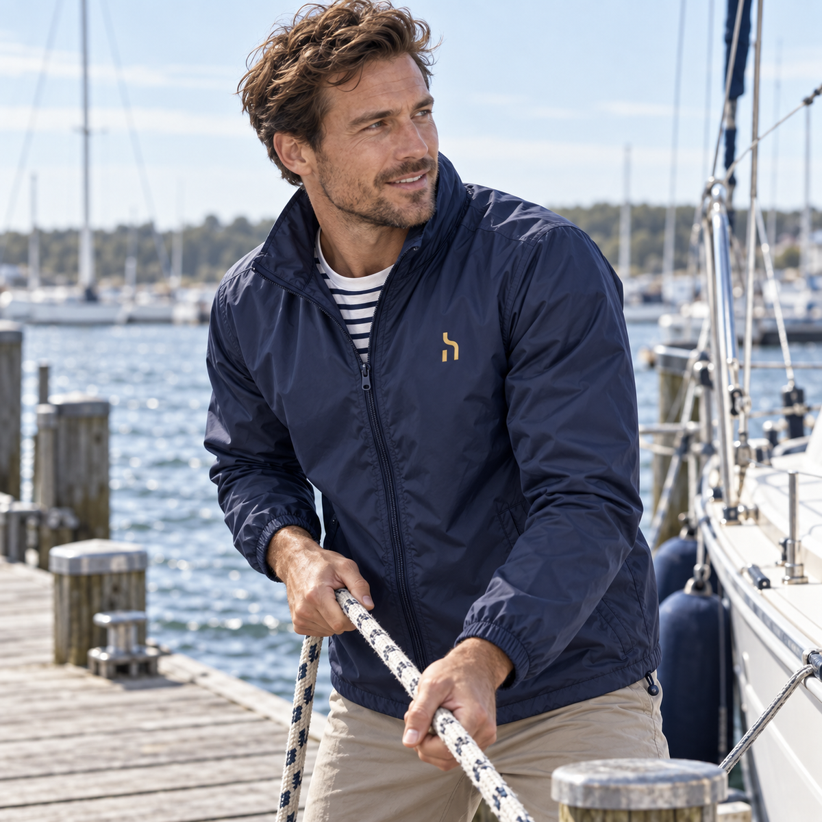 Mann trägt den Navy-Blue-farbenen Haensch Coast Packable Windbreaker mit goldenem Haensch-Zeichen in einem Ostsee-Segelhafen.
