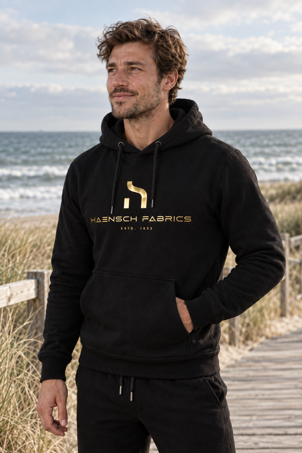Männliches Model trägt den schwarzen Haensch Coast Signature Hoodie mit goldenem Haensch Fabrics Logo an der Ostseeküste