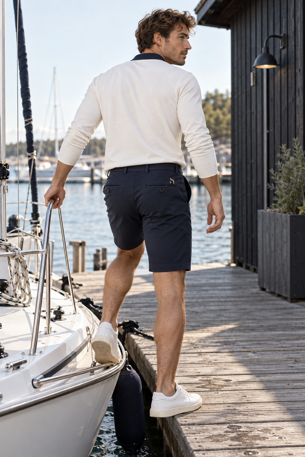 Mann trägt die Haensch Coast Stretch Chino Shorts in Navy mit goldenem Zeichen an einem Ostsee-Segelhafen.