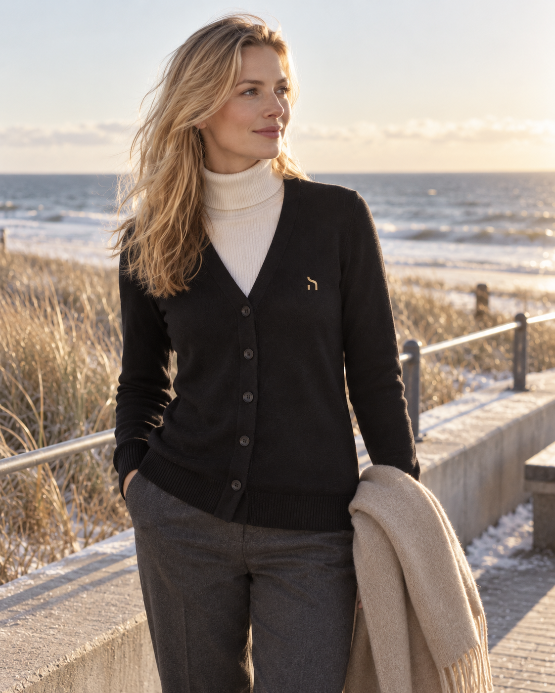 Weibliches Model trägt den Haensch Heritage Supima Cardigan Damen in Winter Black mit goldenem Hänsch-H-Signet und hellem Rollkragen an der winterlichen Ostsee