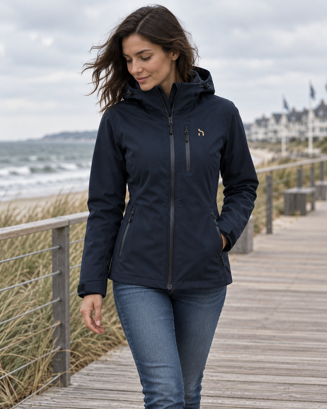 Dunkelhaarige Frau trägt die Haensch Northern All Weather Jacket Damen in Deep Navy beim Spaziergang auf einem Küstenweg an der Ostsee.