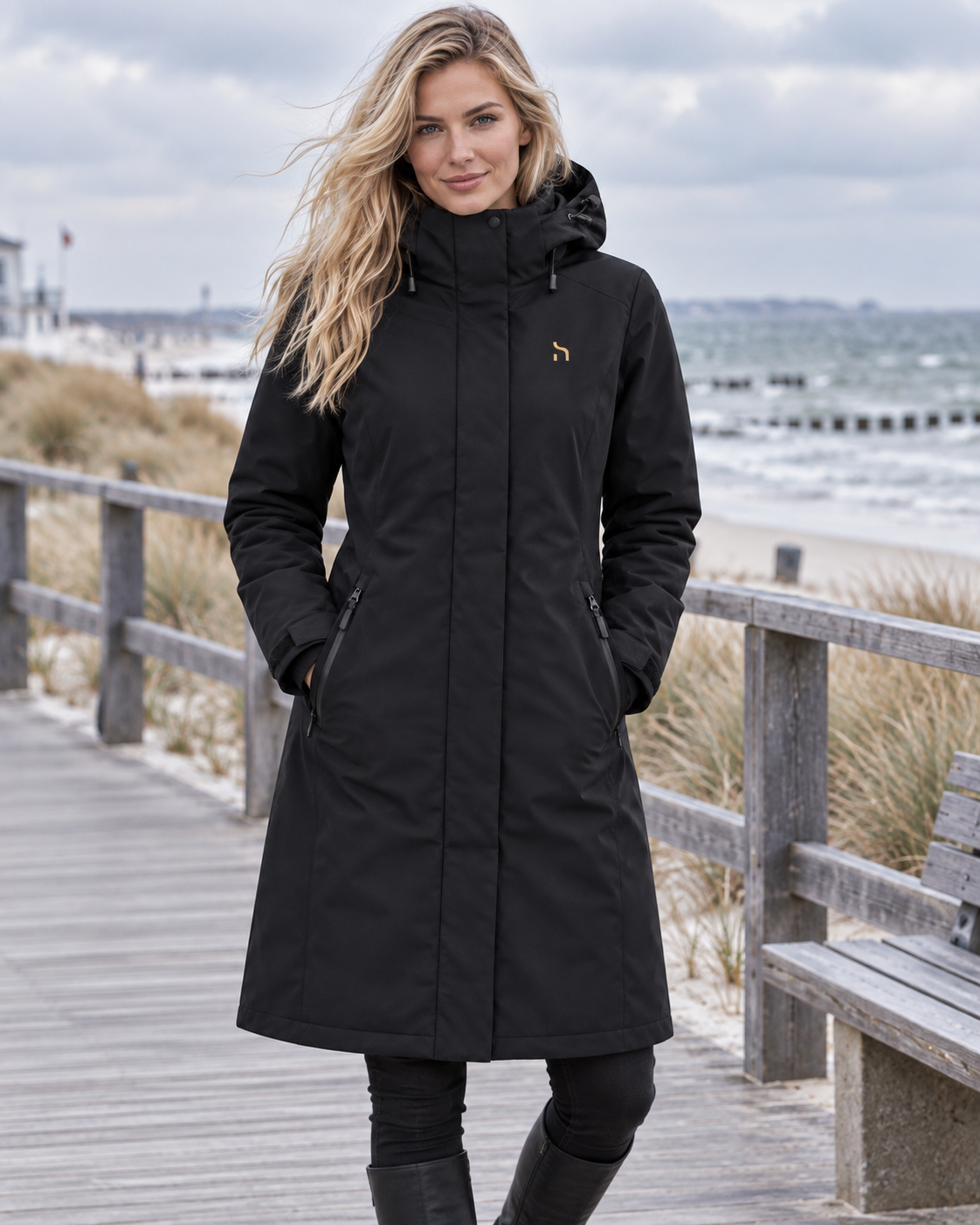Blonde Frau trägt den Haensch Northern All Weather Parka Damen in Winter Black auf einer winterlichen Ostsee-Promenade und blickt direkt in die Kamera.