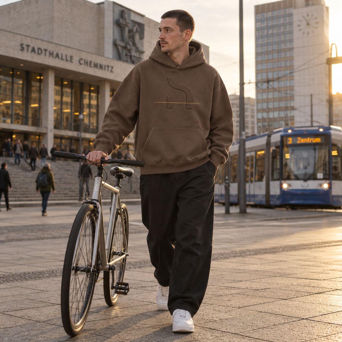 Junger Mann trägt den Haensch Studio Box Hoodie in Bark und schiebt ein Fahrrad vor der Stadthalle Chemnitz.