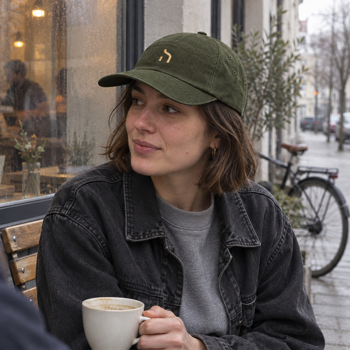 Frau mit olivgrüner Haensch Utility Cap und goldenem Zeichen sitzt mit einer Kaffeetasse vor einem Leipziger Café.